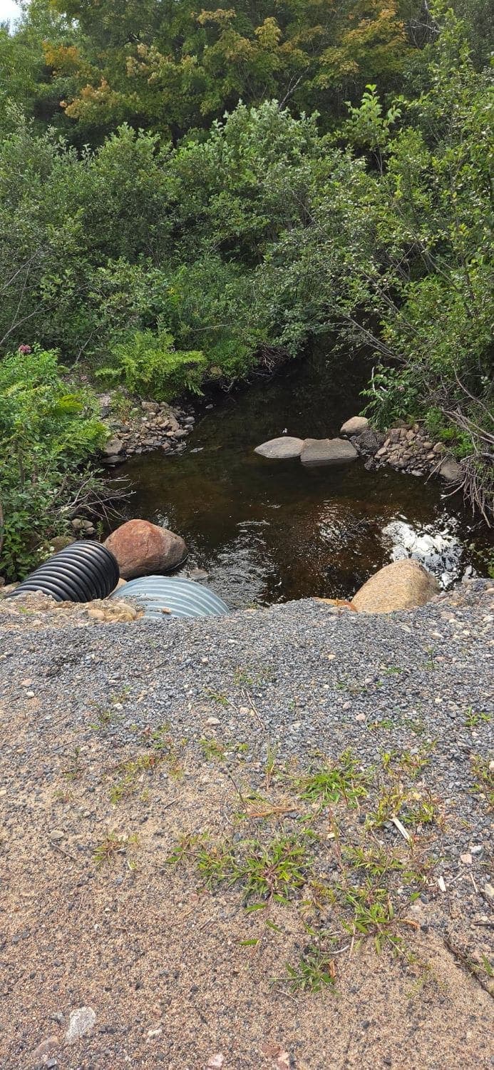 Two culvert pipes under a gravel road where a small creek flows out between boulders